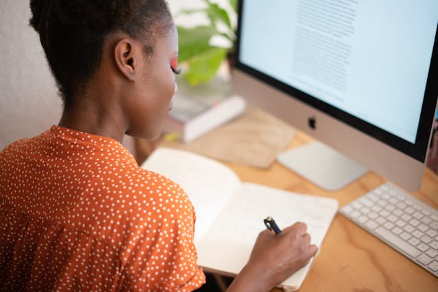 Person writing in a book beside a computer